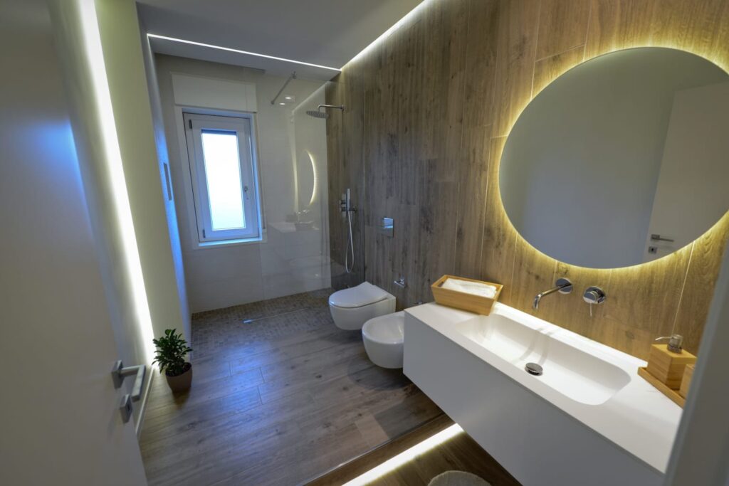 Bagno con rivestimento effetto legno, specchio rotondo retroilluminato e doccia in vetro